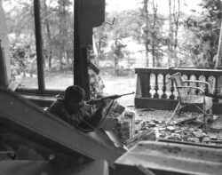 A British Paratrooper taking aim with an American M1 carbine from the first floor balcony of the Hartenstein Hotel in Oosterbeek, near Arnhem in The Netherlands. September 1944. The photograph was taken by Sergeant D M Smith, Army Film & Photographic Unit on Saturday the 23rd of September 1944. Sergeant Dennis Smith, the photographer, wrote: “We have had a very heavy shelling this morning, September 23rd and now the situation is serious. the shelling is hellish. We have been holding out for a week now. The men are tired, weary and food is becoming scarce, and to make matters worse, we are having heavy rain. If we are not relieved soon, then the men will just drop from sheer exhaustion”.
The British 1st Airborne Division headquarters had been established in the Hotel during ‘Operation Market Garden’ and it is now the Airborne Museum ‘Hartenstein’.