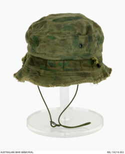 Australian bush hat : Trooper D R Barnby, 2 Squadron, Special Air Service Regiment. Description: Modified Australian Army issue cotton patrol 'giggle' hat with shortened brim and green nylon chin strap attached. The nylon chin strap is attached to the hat by a pair of holes made into the side of the hat with a knot keeping it in place on either side. An adjustable plastic toggle allows the wearer to tighten or loosen the chin strap. A pair of circular metal ventilation holes are on both sides of the crown. A mixture of faded green and black paint has been randomly applied to the exterior as a means of camouflaging the hat. History / Summary: The Special Air Service Regiment (SASR) in Vietnam were well known for modifying issued equipment for their own unique purposes. This hat is an example of this adaptive attitude. The brims of many SASR hats were removed to allow a better field of vision for the wearer, and the added chin strap ensured the hat would not be lost on patrol or in transport. AWM Accession Number: REL/14214.002 SASR Don Barnby Vietnam SAS